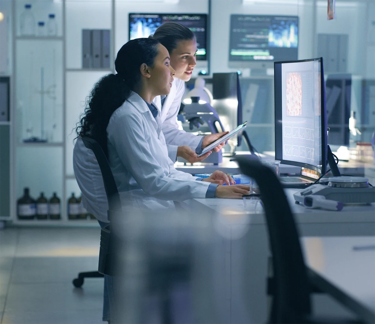 Two female scientists in lab coats analyzing data on a computer screen in a modern lab.