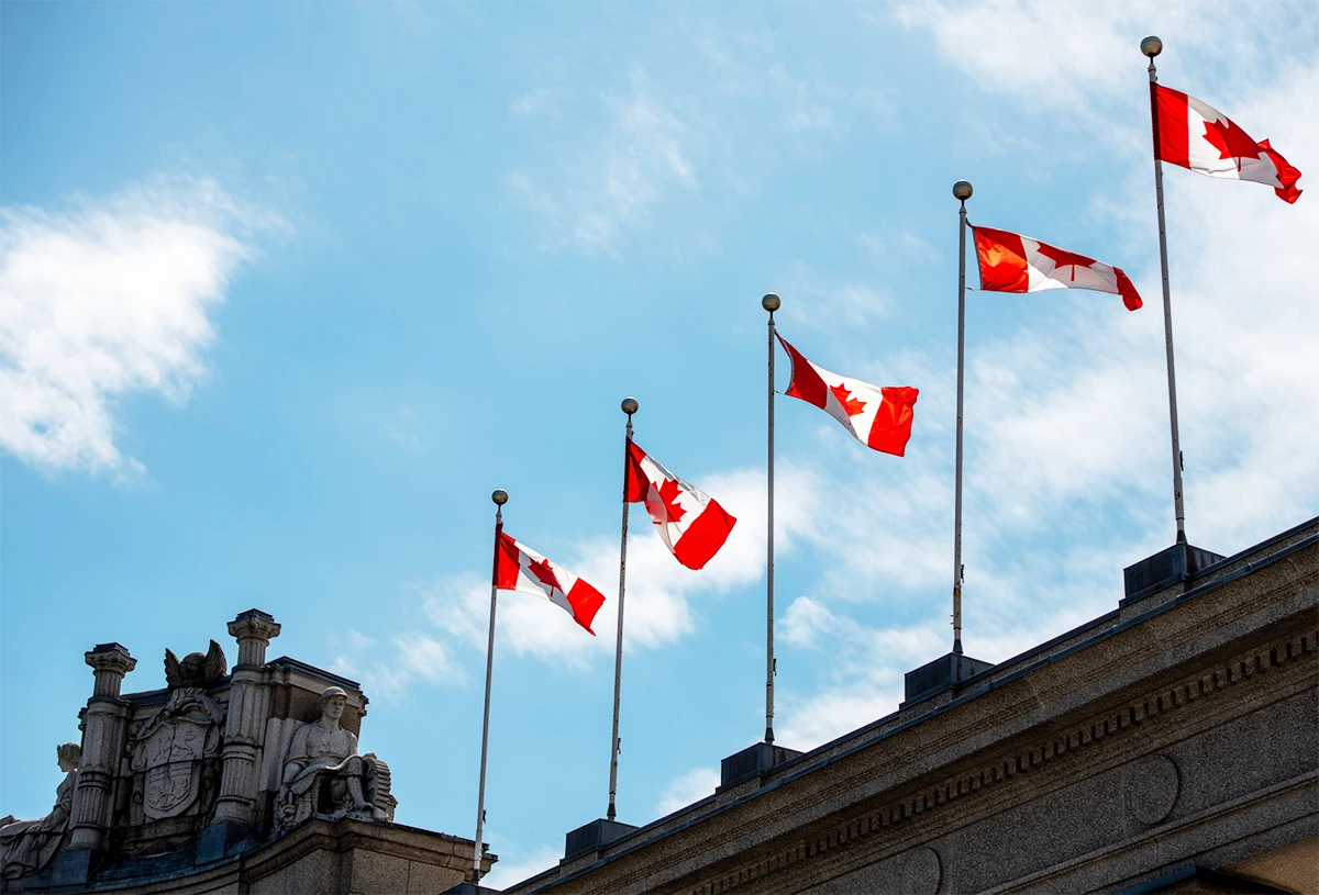 Five Canadian flags on poles atop a stone building against a partly cloudy blue sky.