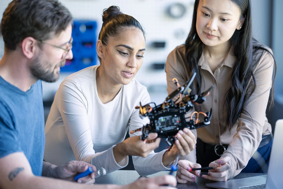 Three people examining and discussing a robotic crab device in a workshop setting.