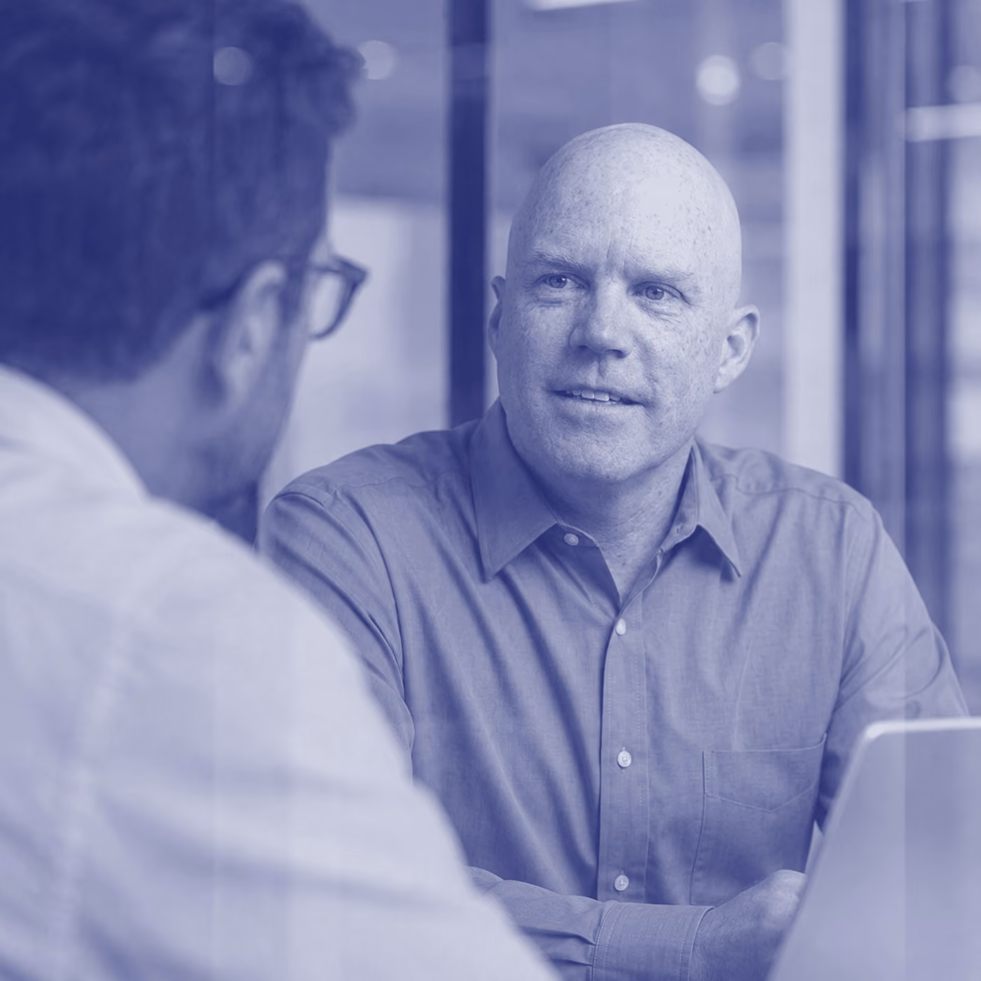 Bald man in a button-up shirt in conversation with a colleague in an office, in blue duotone