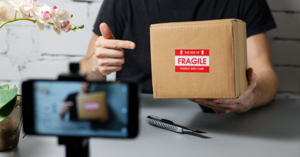 Smartphone on a stand recording a livestream while a person holds up a cardboard box with a red "Fragile" label in the background.