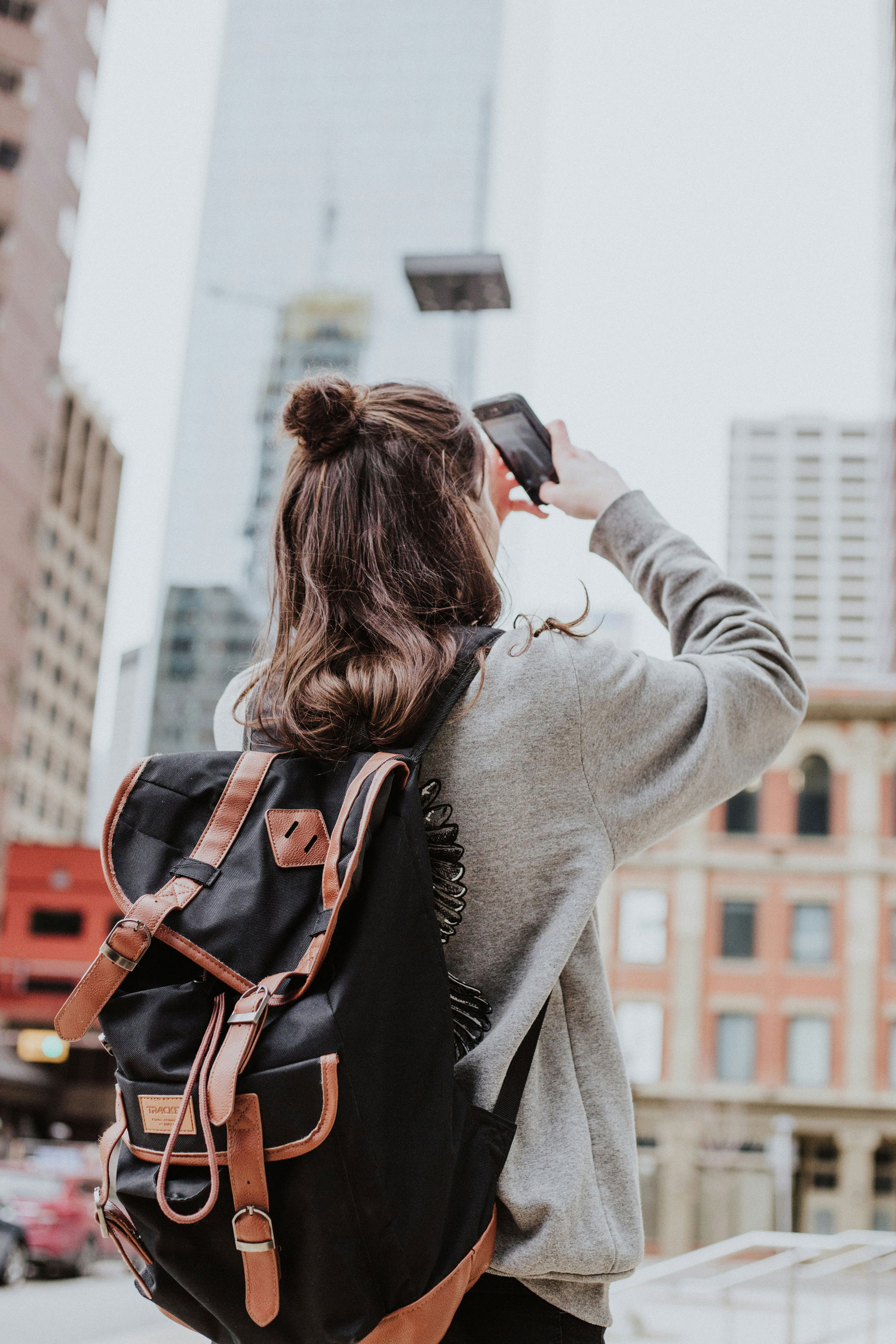 Person with a black backpack and gray sweater taking a photo with a smartphone in a city.