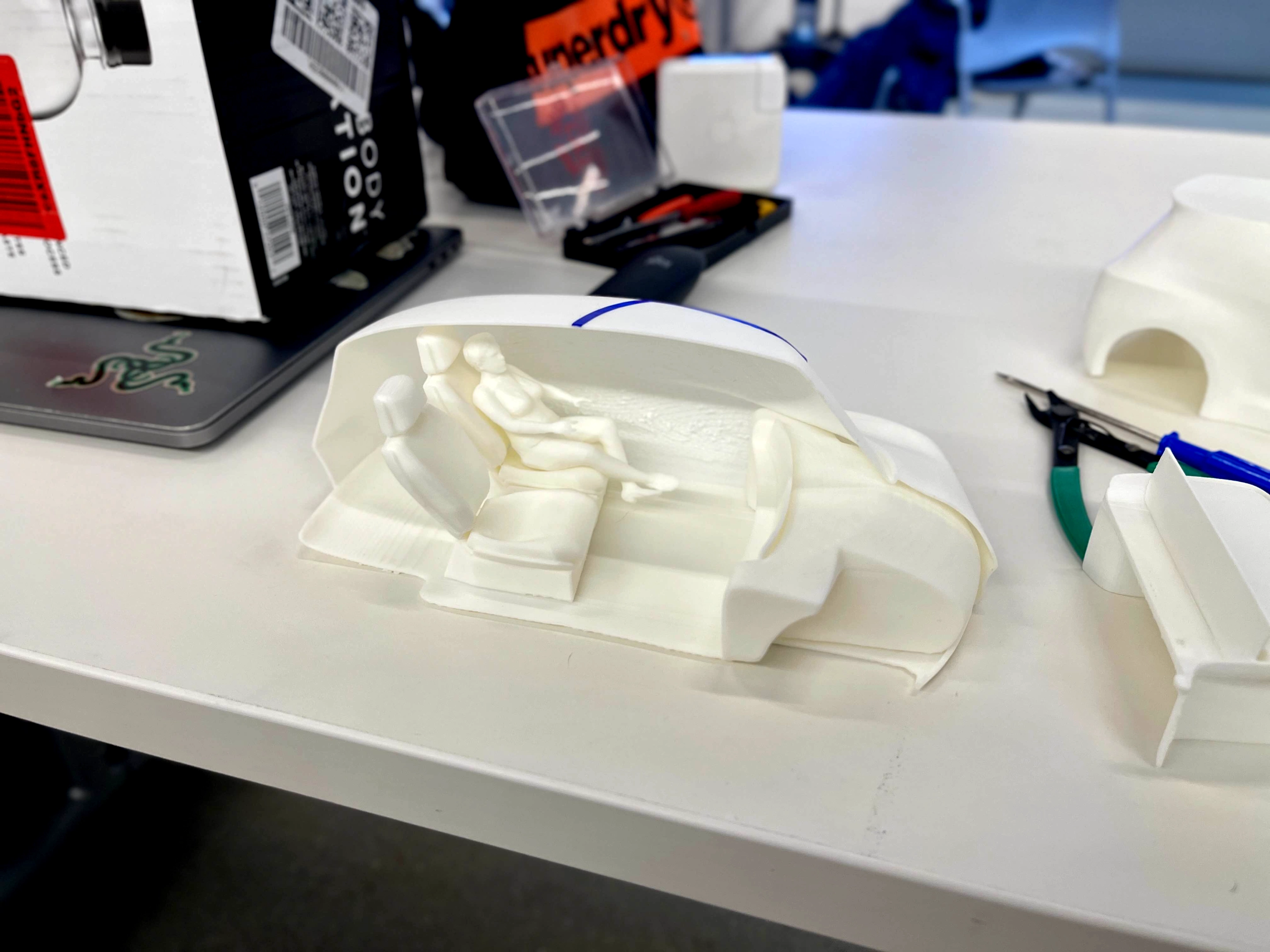 White 3D-printed car interior model with seats and a figure seated inside on a white table.