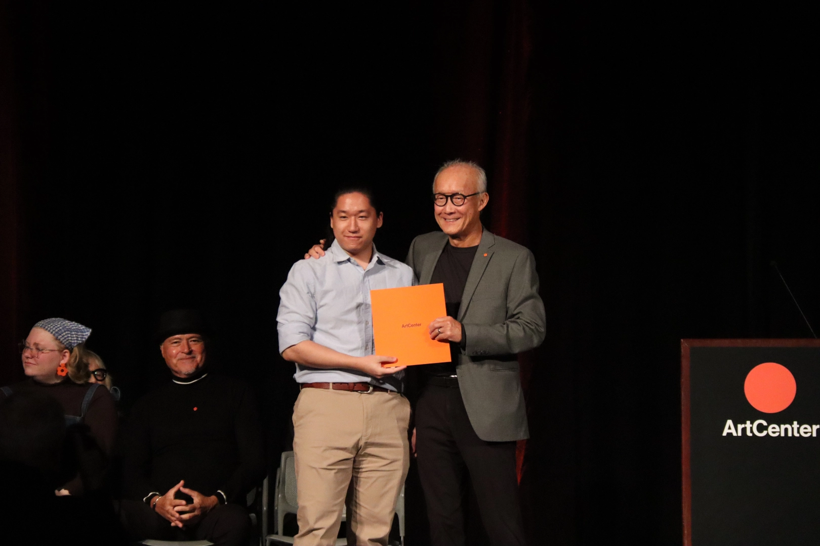 Two men smiling on stage holding an orange folder with ArtCenter logo, others seated nearby.