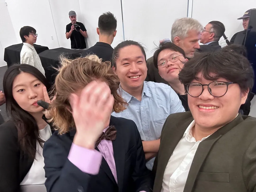 Group selfie of six people smiling, one adjusting hair, in a casual indoor setting.