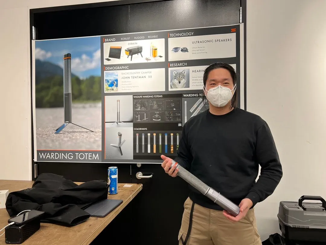 Person with mask holding a metallic device in front of a board detailing the Warding Totem product.