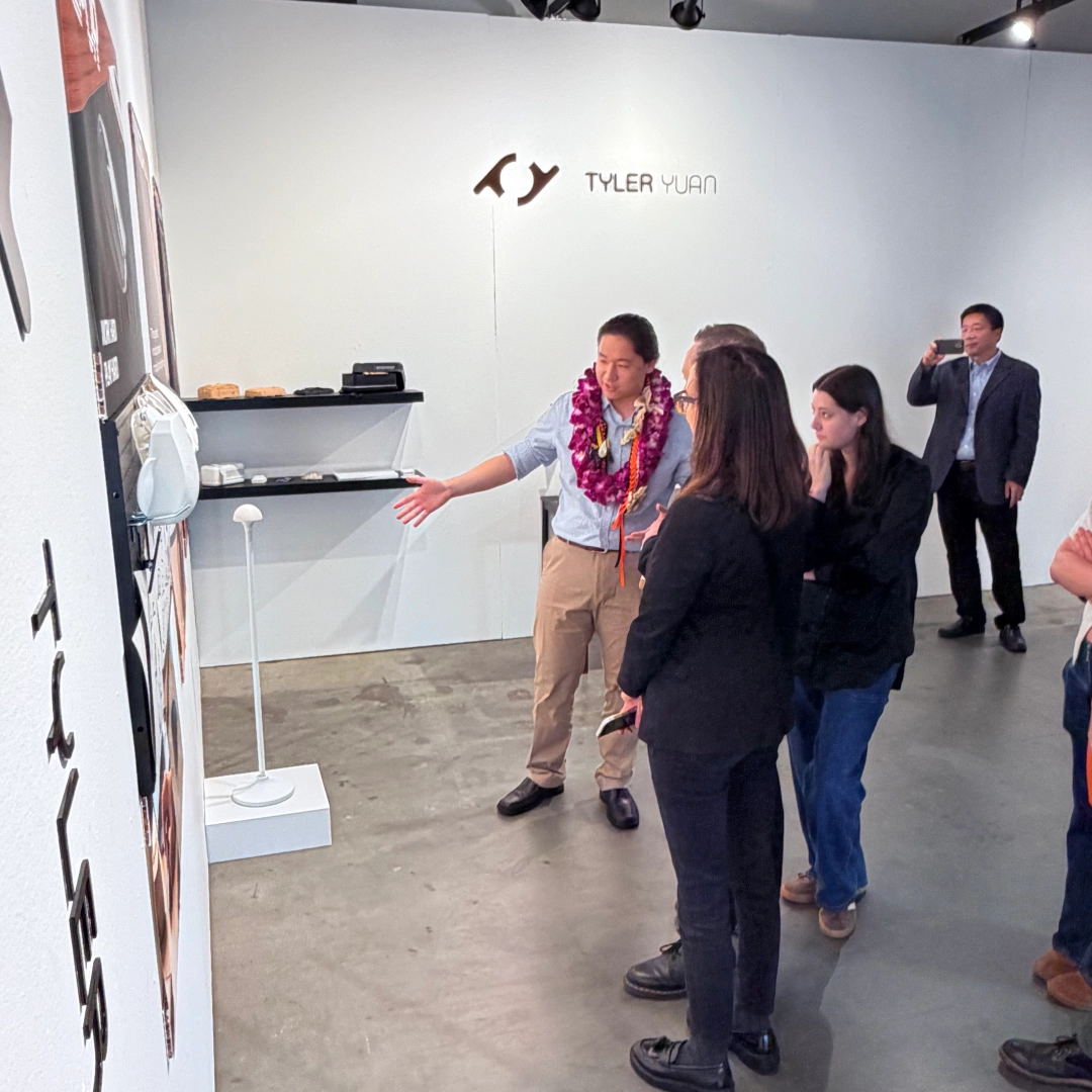 Man with lei gestures while explaining artwork to three people in a gallery with 'Tyler Yuan' on the wall.