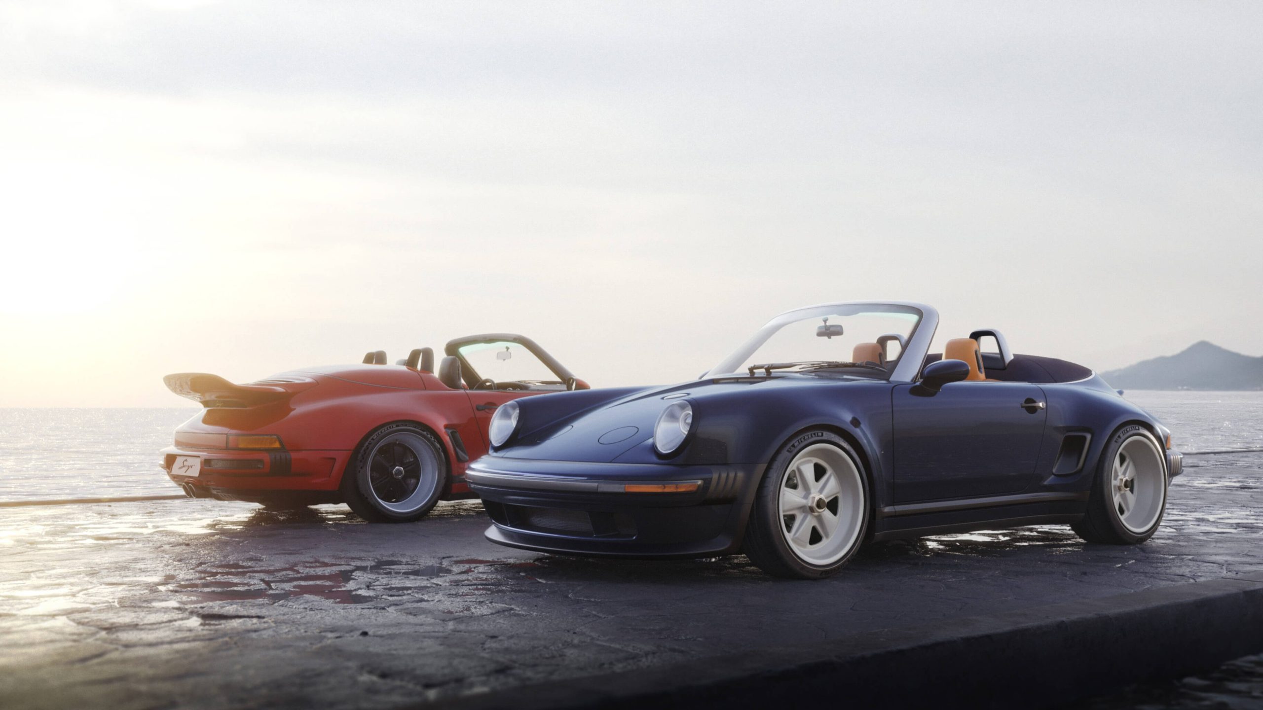 Two classic convertible sports cars, one navy blue and one red, parked on a stone pier by the ocean.