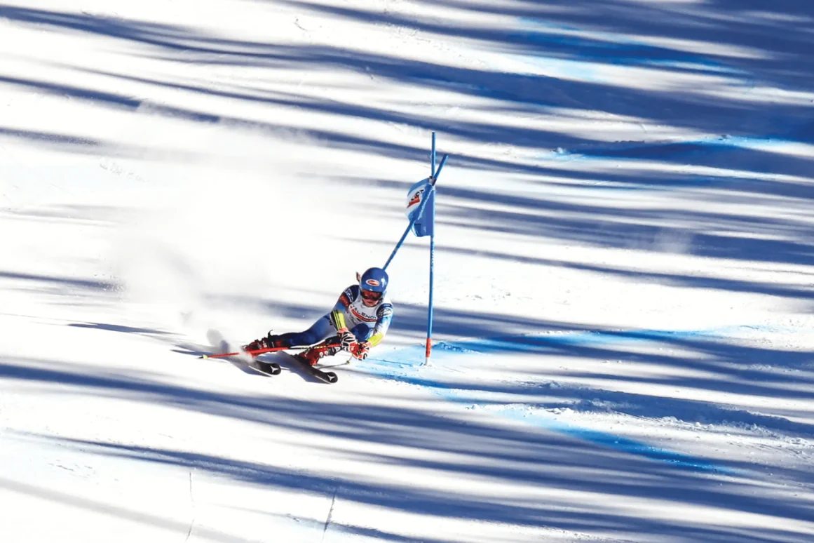 Alpine skier racing downhill around a blue gate on a snowy slope casting striped shadows.