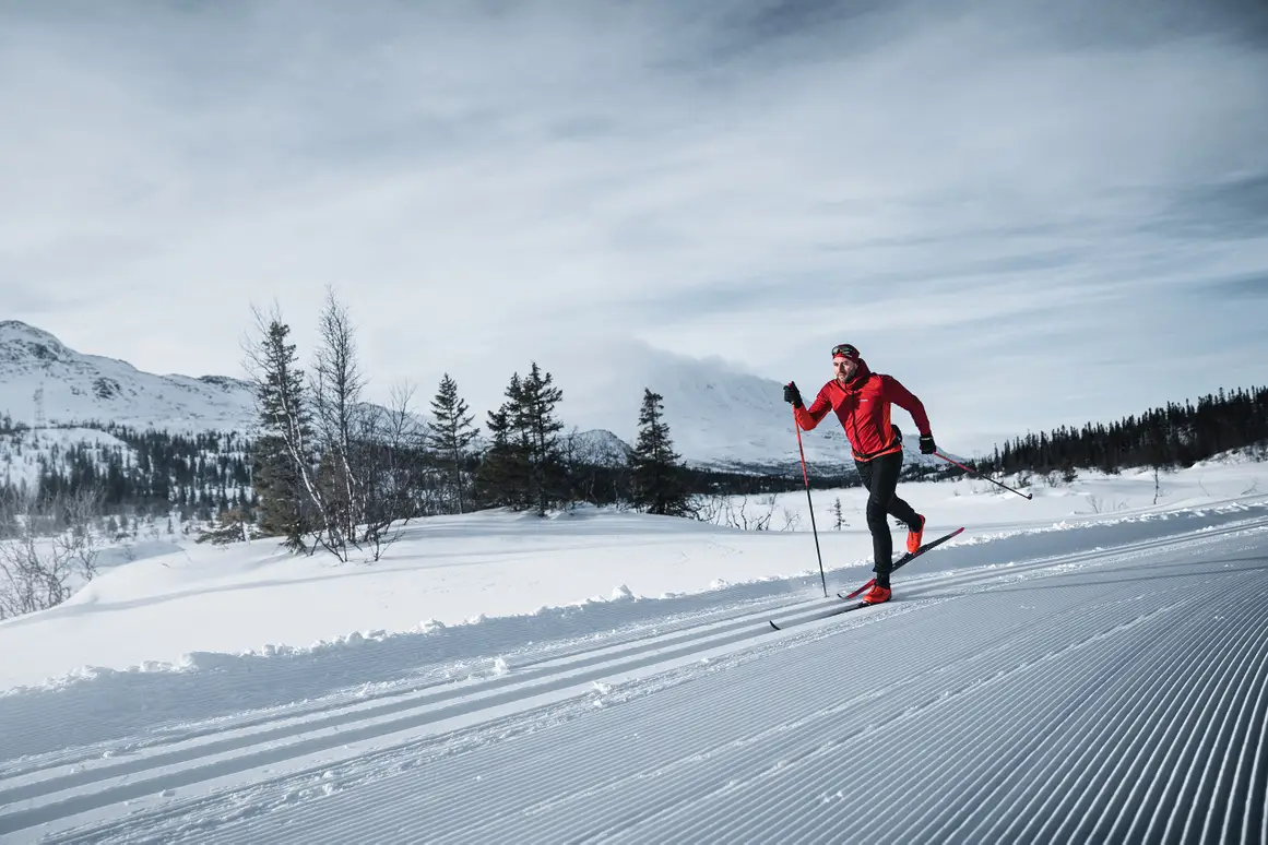 Man skiing on a groomed snowy trail with snow-covered mountains and trees in the background.