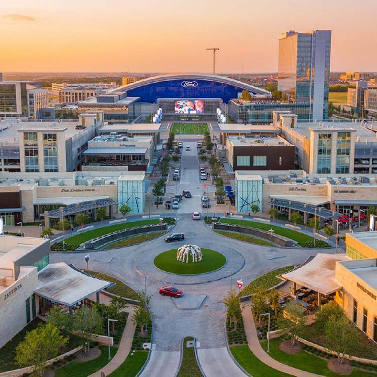 A wide street with shops, a fountain, and a stadium in Frisco at sunset.