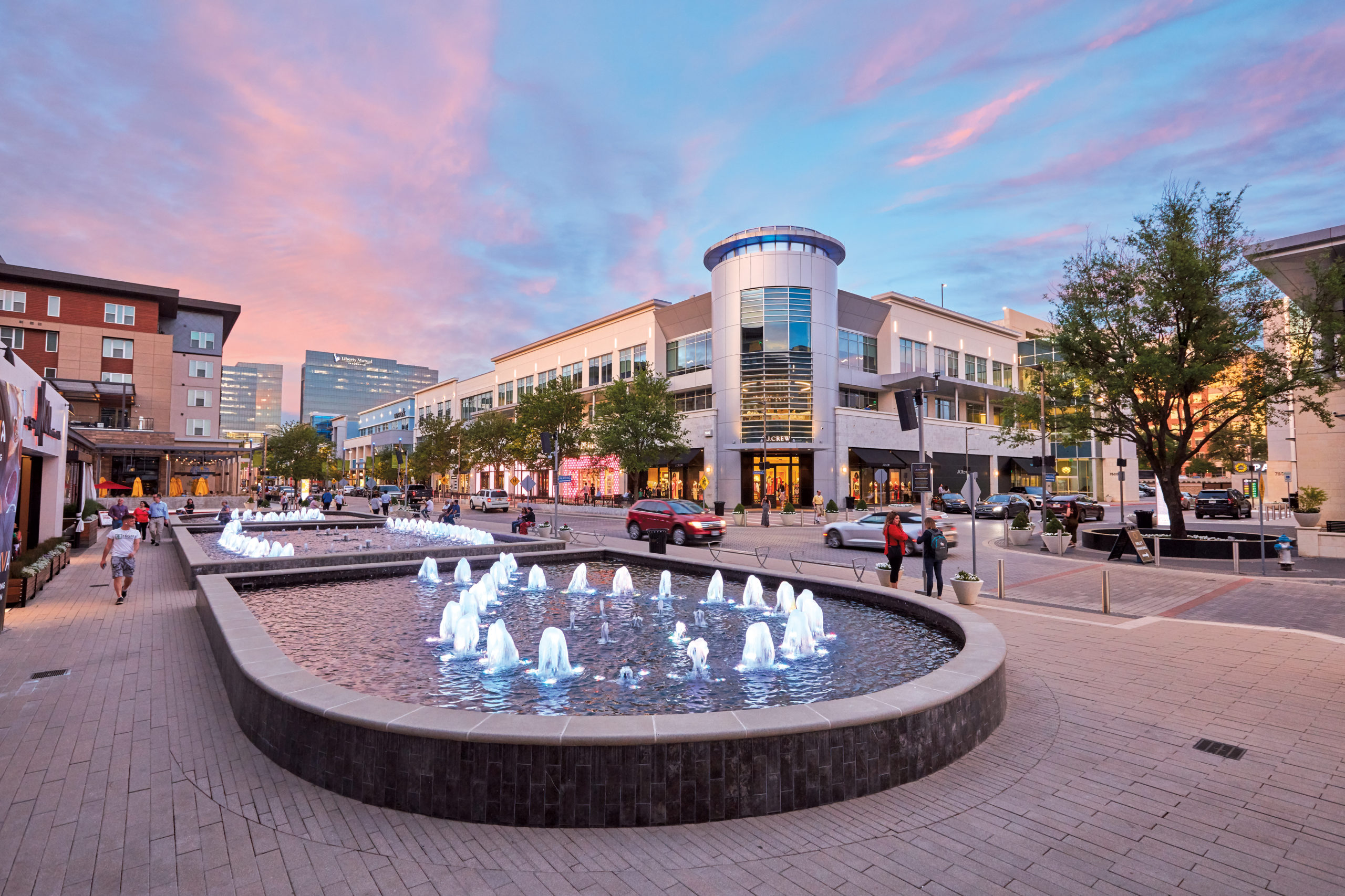 A fountain and shops in downtown Plano at sunset with people walking nearby.