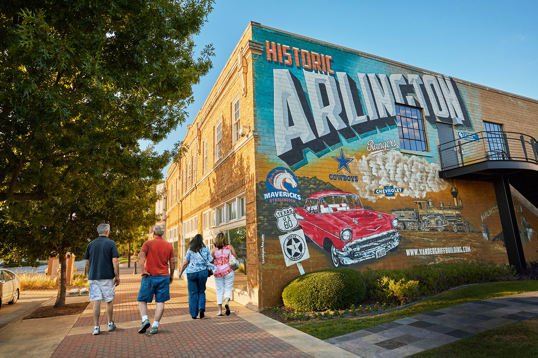 Four people walk past a large Historic Arlington mural painted on a brick building.