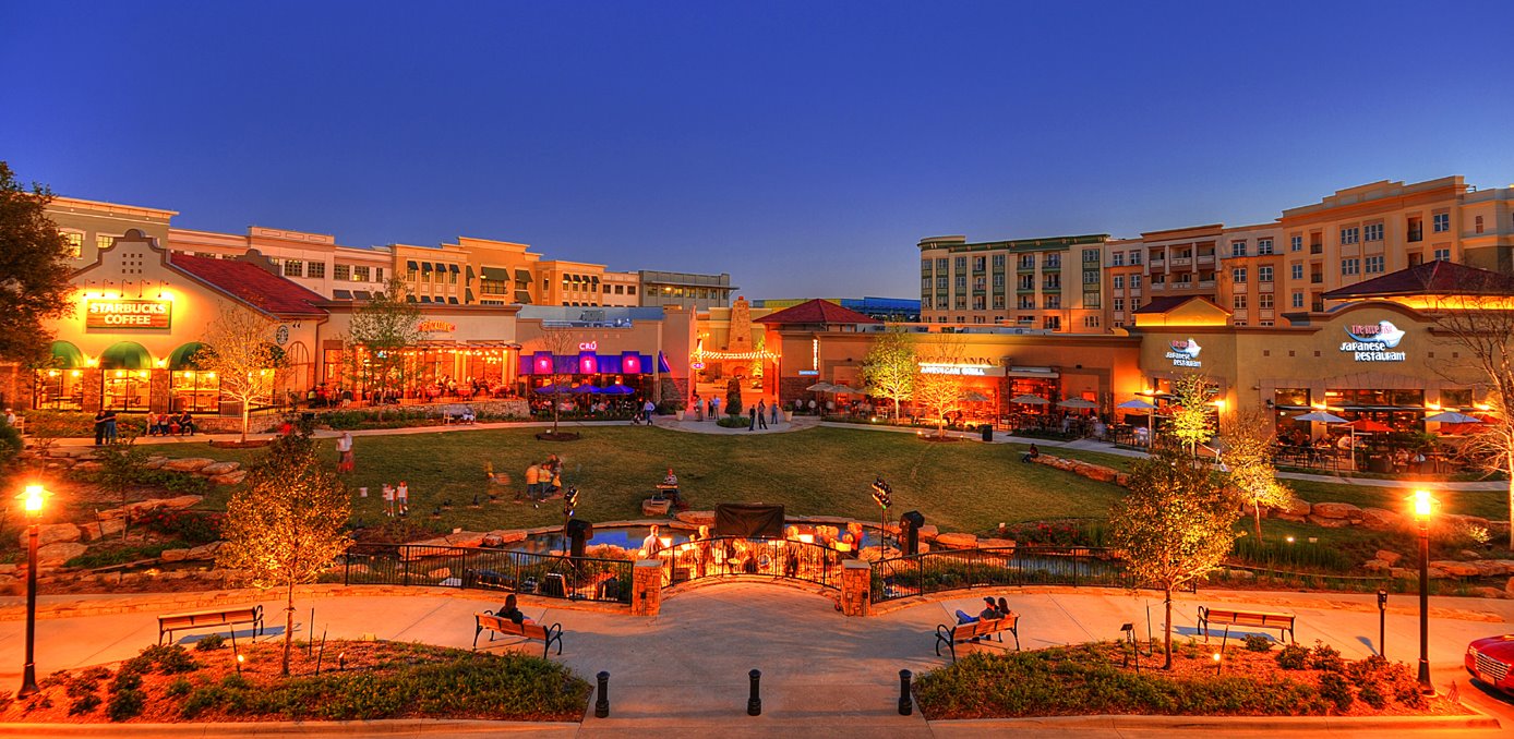 Shops and a green lawn in Allen at night with people sitting on benches.