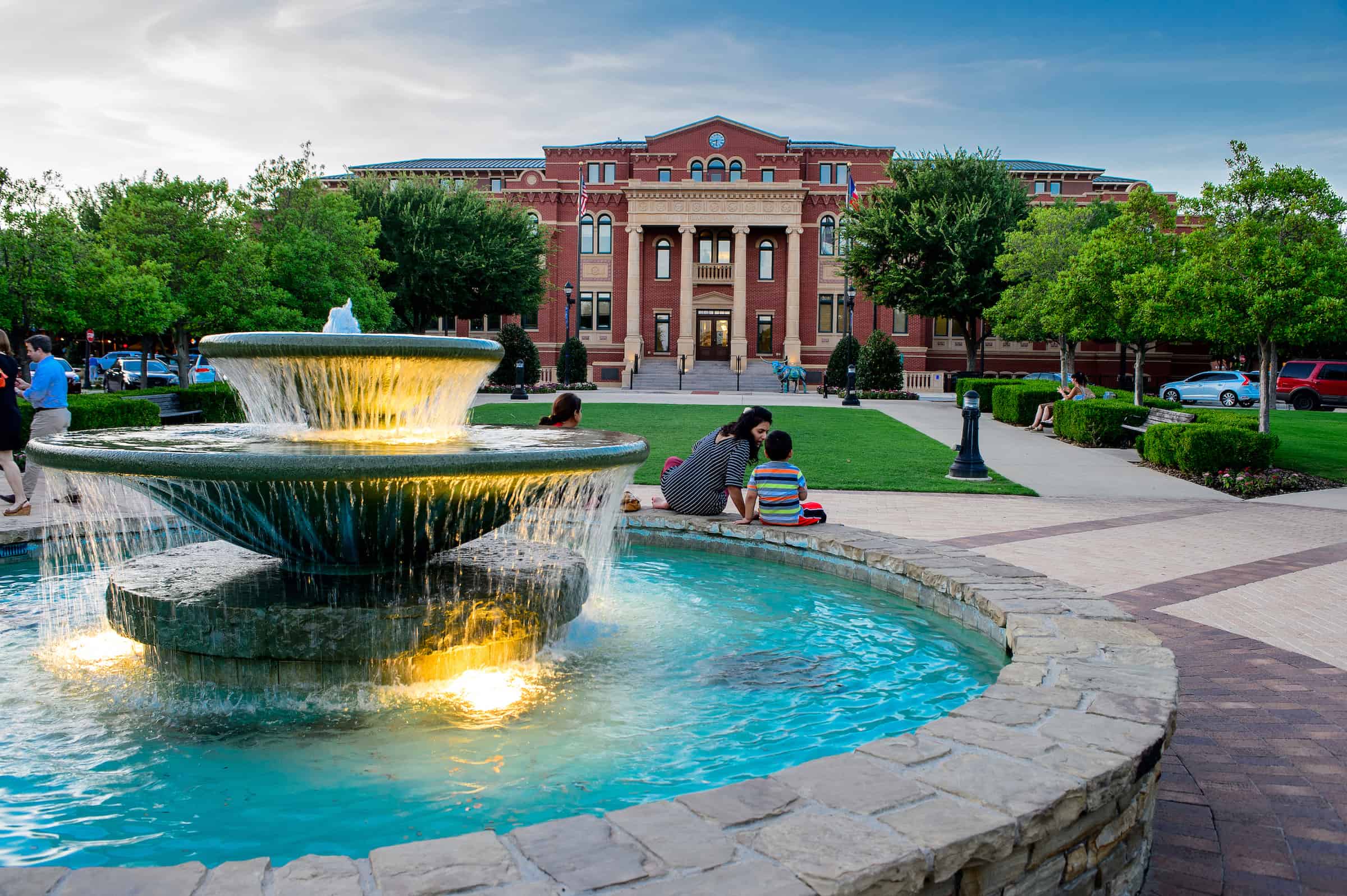 A fountain in Southlake Town Square with families sitting nearby and a brick town hall behind.