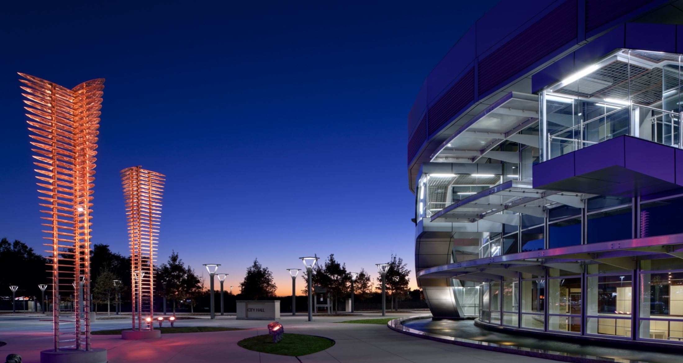 A modern glass building in Mesquite lit up at dusk with tall lighted sculptures nearby.