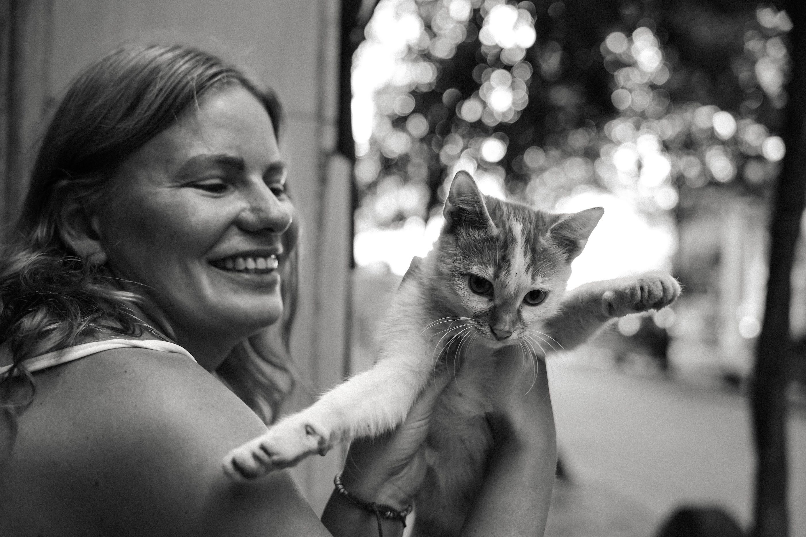 Smiling woman holding a cat with outstretched front legs against a blurred outdoor background.