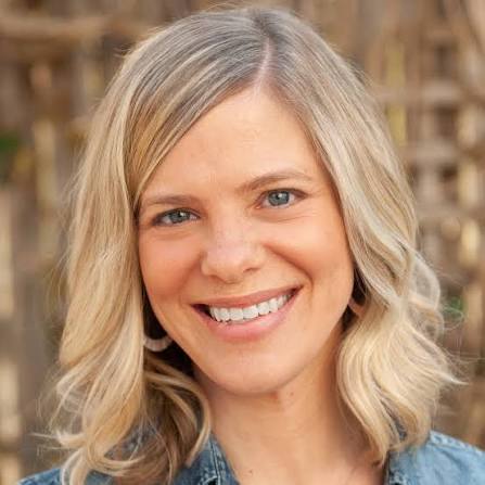 Smiling woman with blonde wavy hair and blue eyes wearing earrings and a denim shirt.