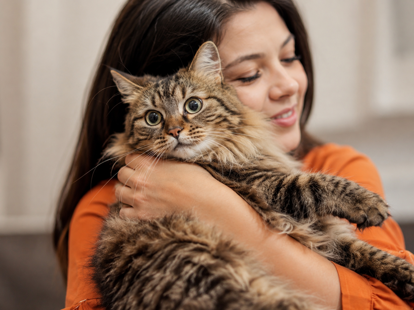 Woman in orange shirt hugging a large fluffy tabby cat with wide green eyes indoors.