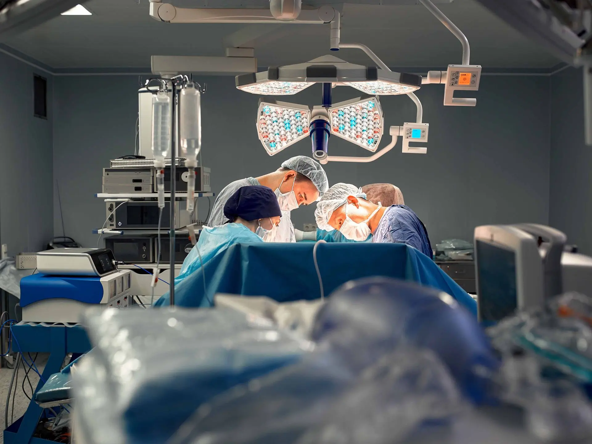 Three surgeons wearing masks and operating under surgical lights in an operating room.
