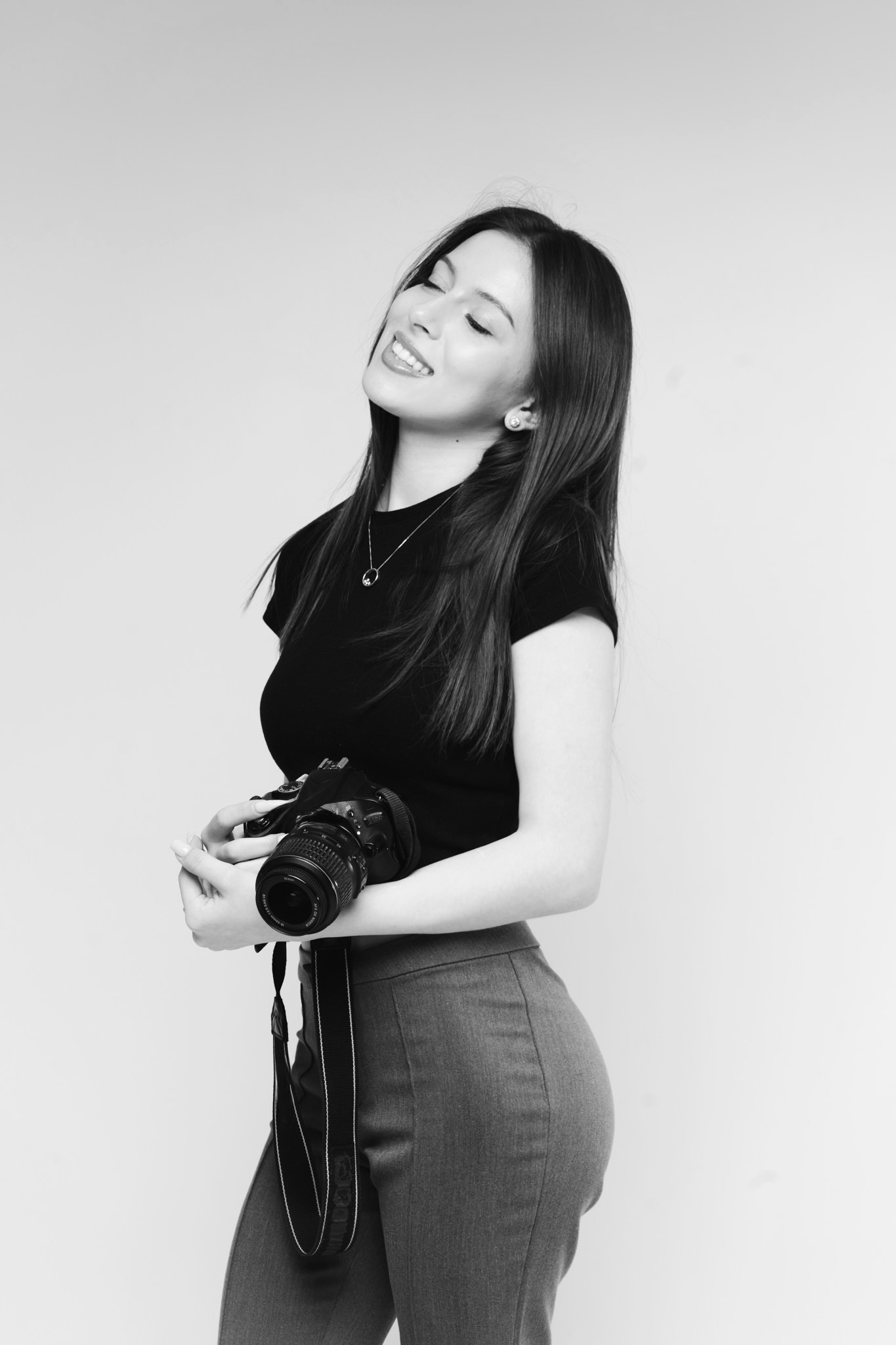 Young woman smiling with closed eyes, holding a DSLR camera with strap, standing against a plain background.