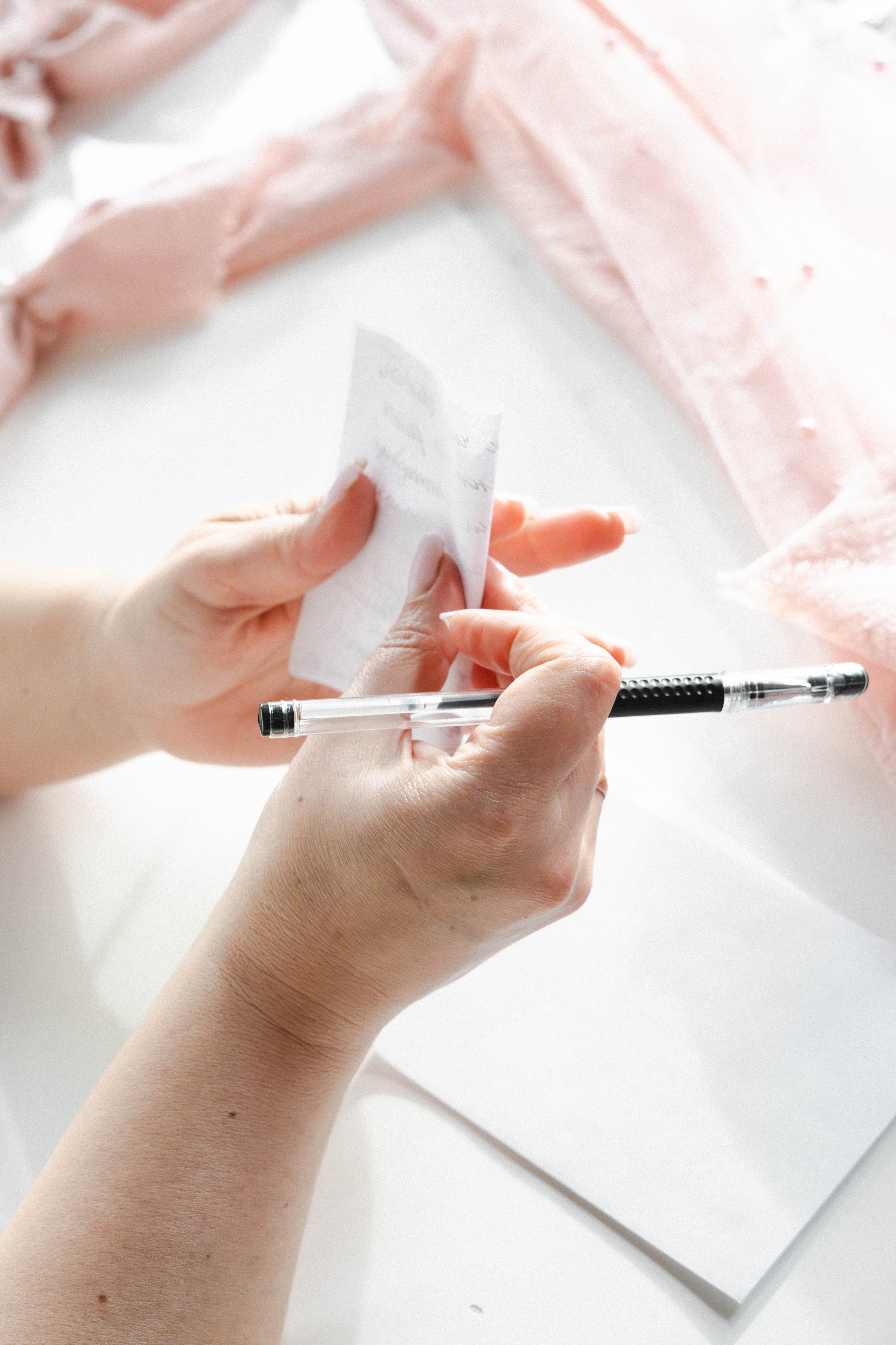 Close-up of hands holding a small piece of paper and a pen, with a pink knitted cloth and a blank notepad on a white surface in the background.