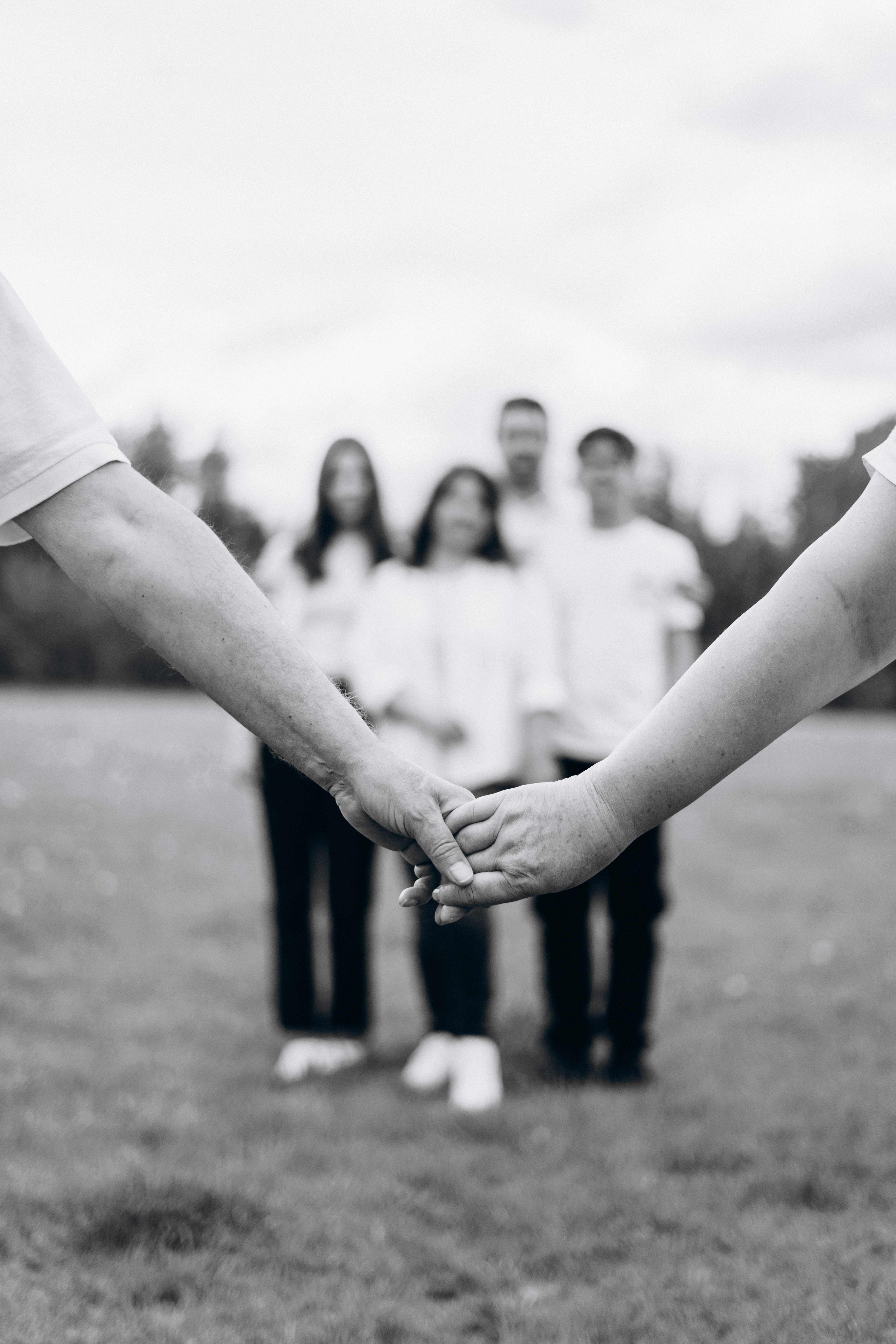 Black-and-white photo of two people in the foreground holding hands, with a blurred group of four people standing together in the background on a grassy field.
