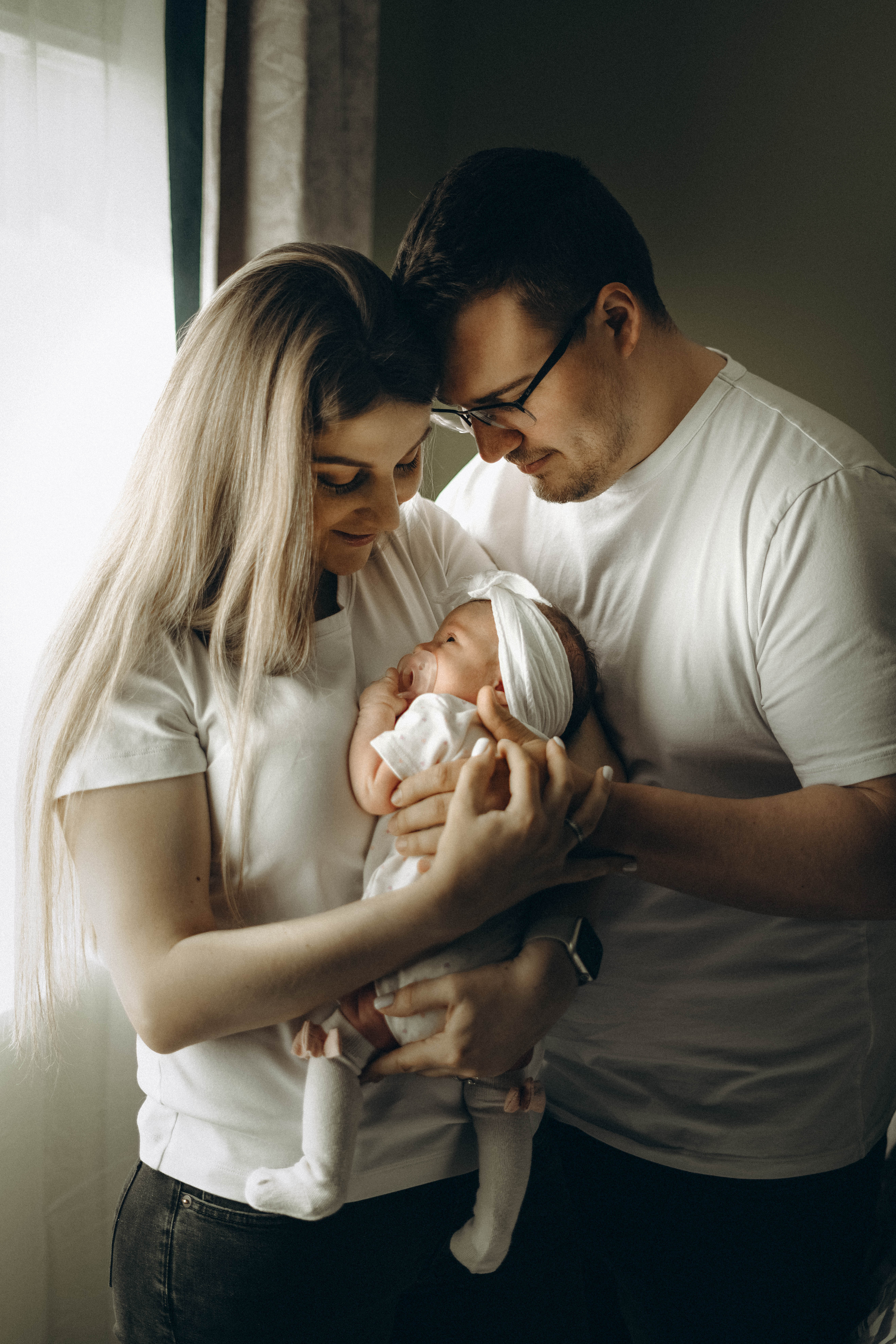 A couple holds a baby together near a window, both looking affectionately at the infant wearing a headband and white outfit with socks.