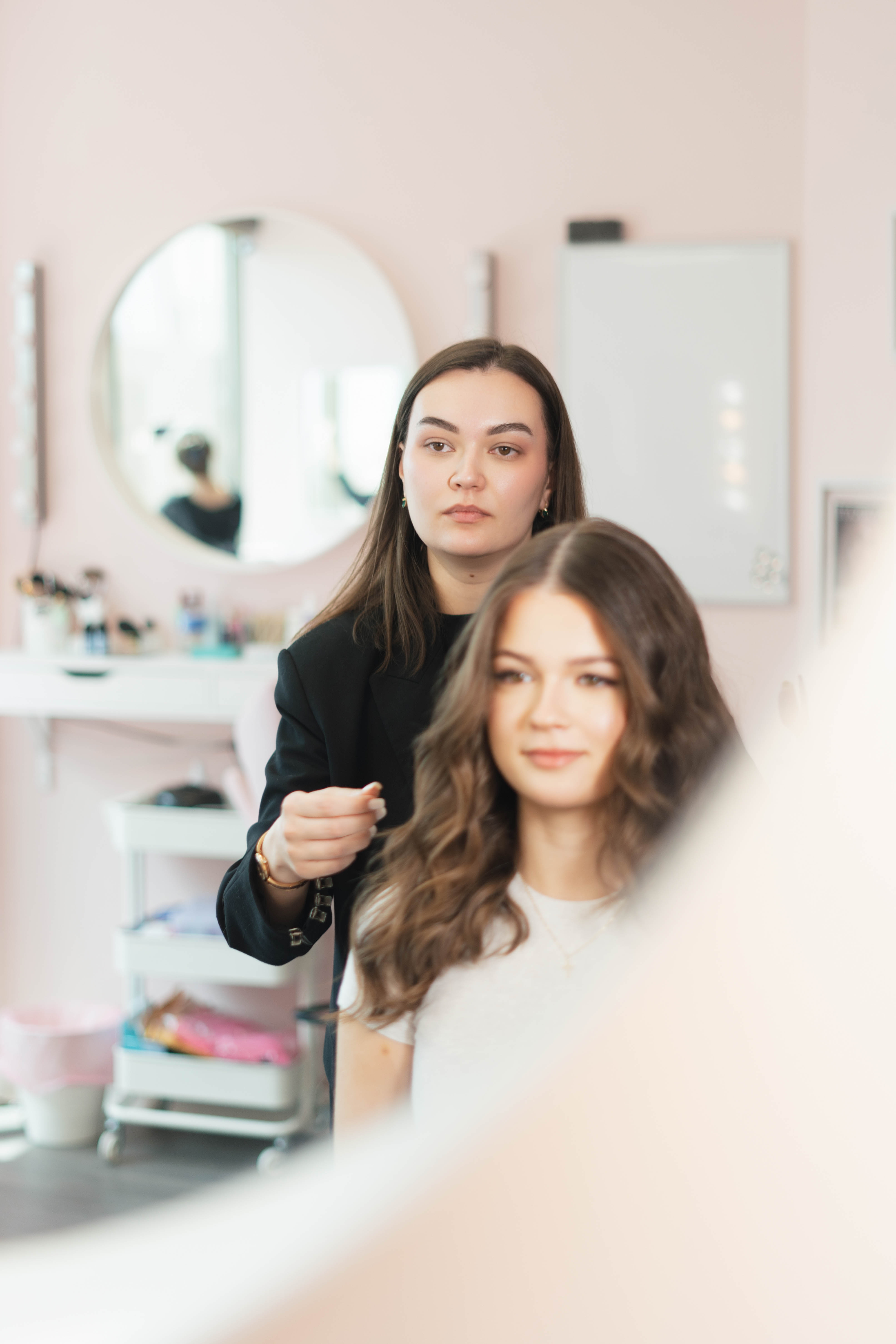 A hairstylist with dark hair in a black jacket styling the hair of a smiling woman with long wavy brown hair in a light-colored top, in a salon with a round mirror and makeup tools in the background.