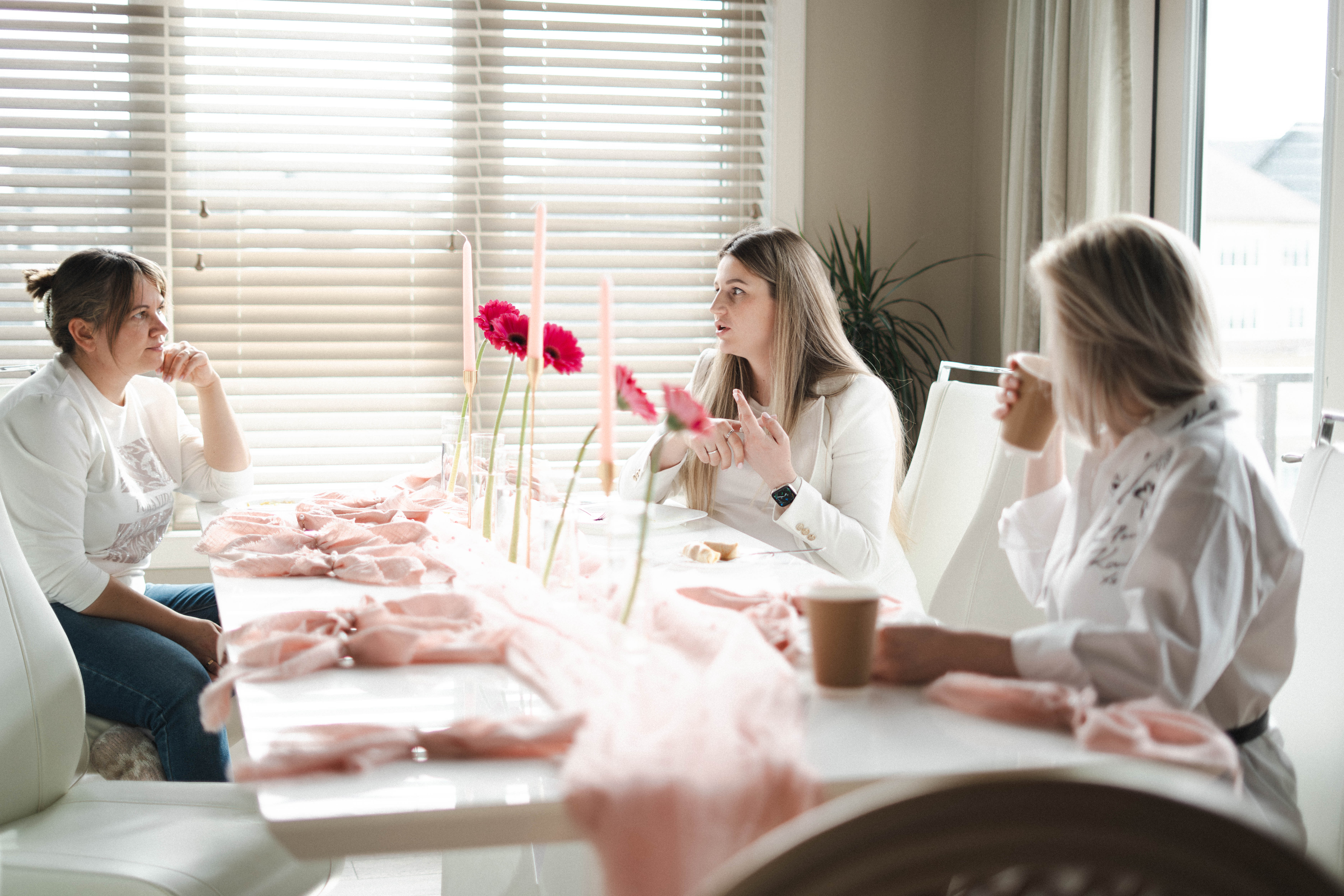 Three women seated around a white dining table with pink candles and flowers, engaged in conversation and drinking from cups, in a room with large windows and blinds.