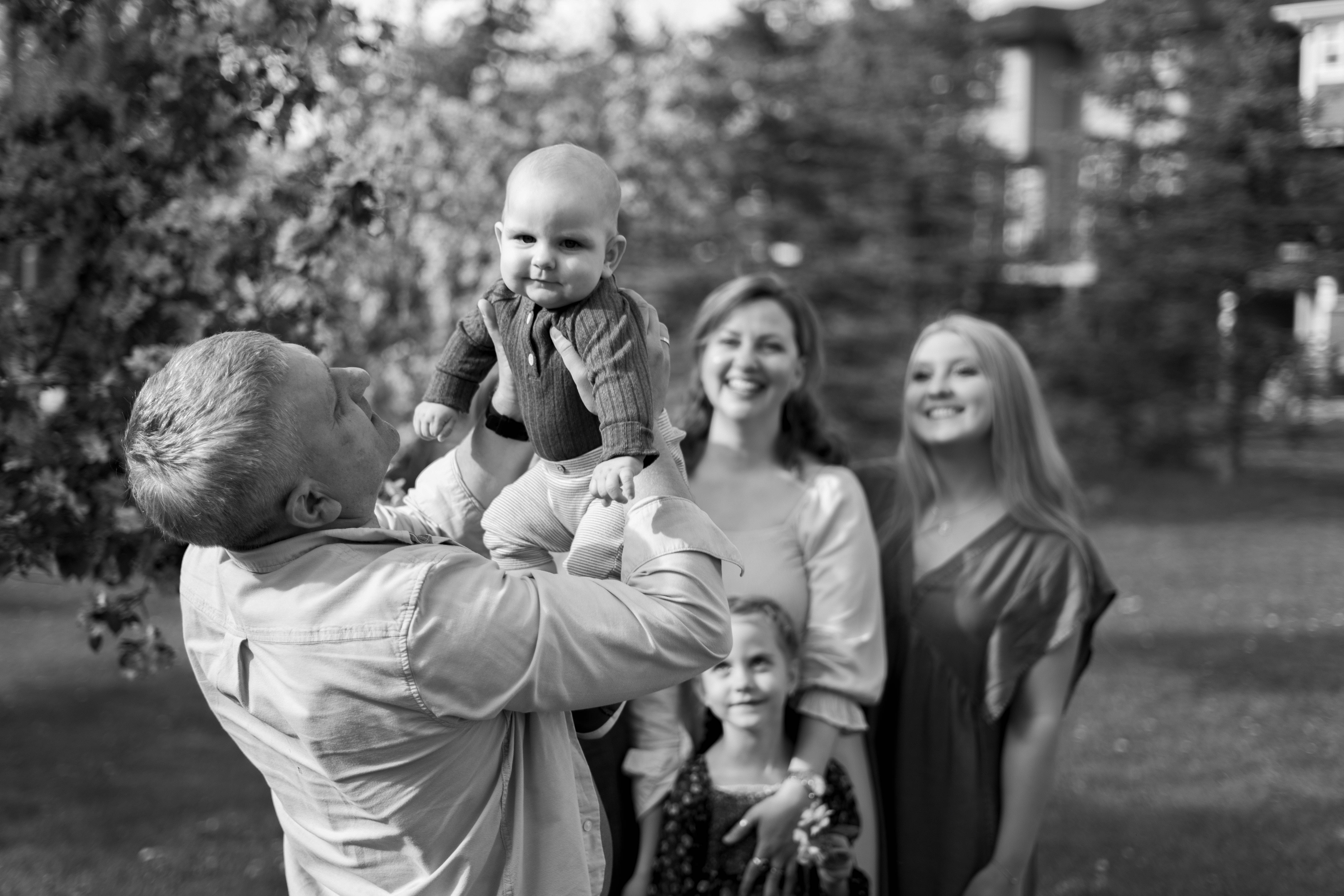 Man lifting a baby in the air outdoors with a smiling woman, young girl, and another woman in the background.