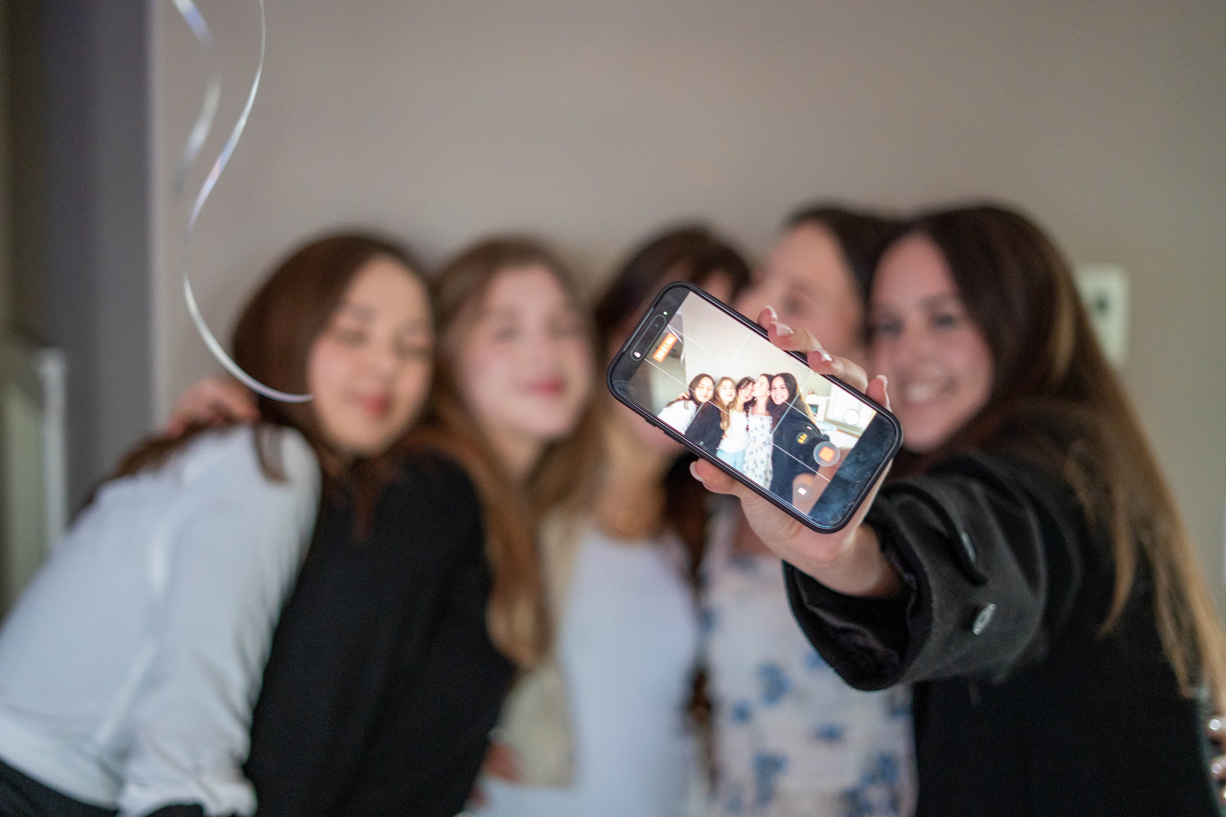 A group of five young women smiling and posing closely together while taking a selfie with a smartphone held in the foreground, which shows their clear image.