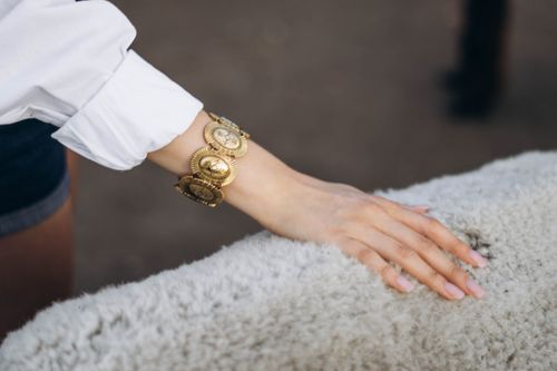 Close-up of a woman's hand with pink nails wearing a large gold bracelet touching soft, fluffy white wool.