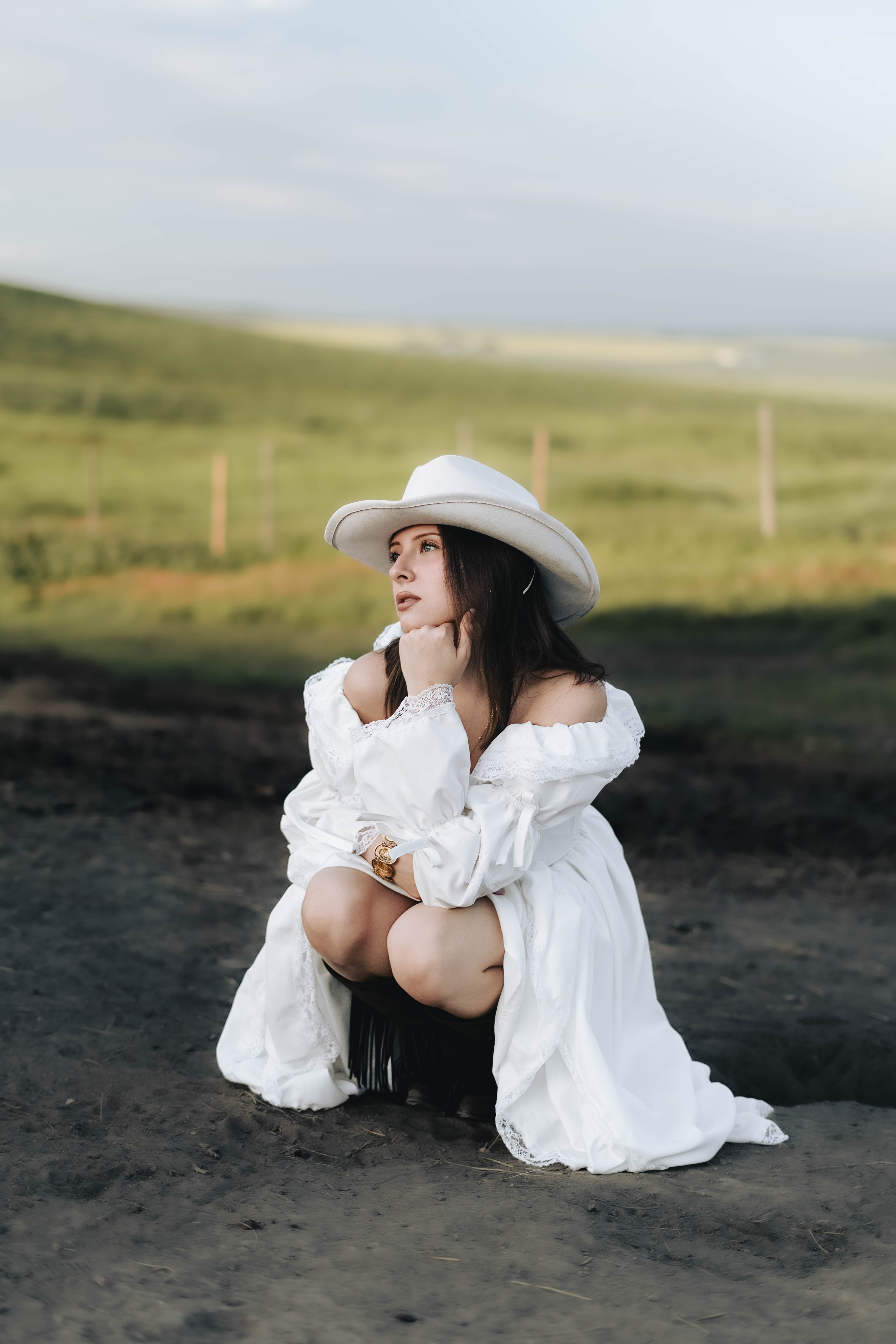 Woman in a white off-shoulder dress and white cowboy hat crouching on a dirt ground with a green field background.