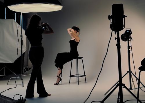 Photographer taking pictures of a woman posing on a stool in a studio with professional lighting equipment.