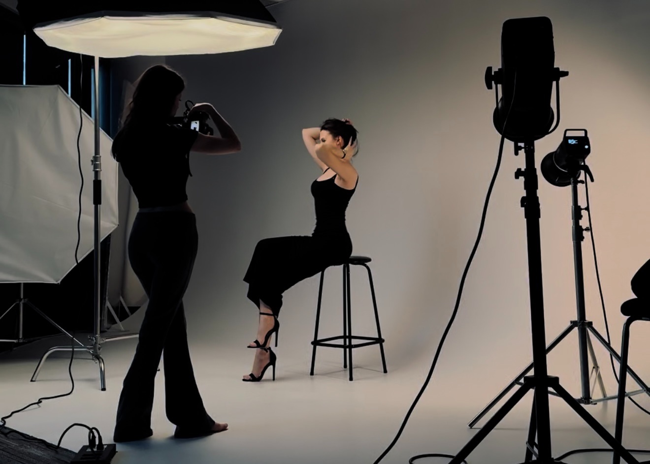 Photographer taking pictures of a woman posing on a stool in a studio with professional lighting equipment.