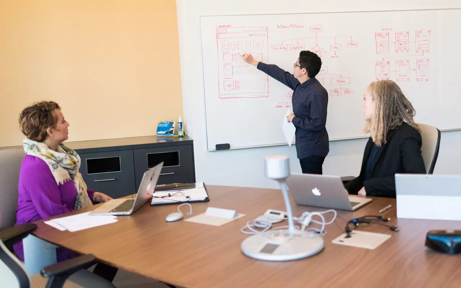 Colleagues in a meeting room reviewing a wireframe sketch on a whiteboard