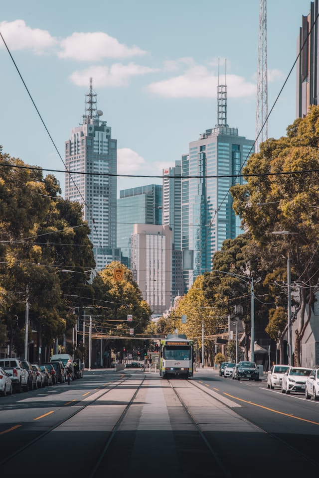City street with tram tracks, a tram, parked cars, green trees, and tall modern skyscrapers in the background.