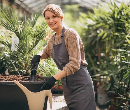 Woman wearing gloves and apron gardening with small shovel near plants in greenhouse.