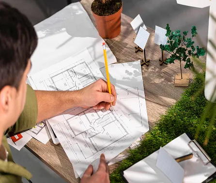 Person drawing on architectural plans with a pencil beside model trees and a small potted plant.