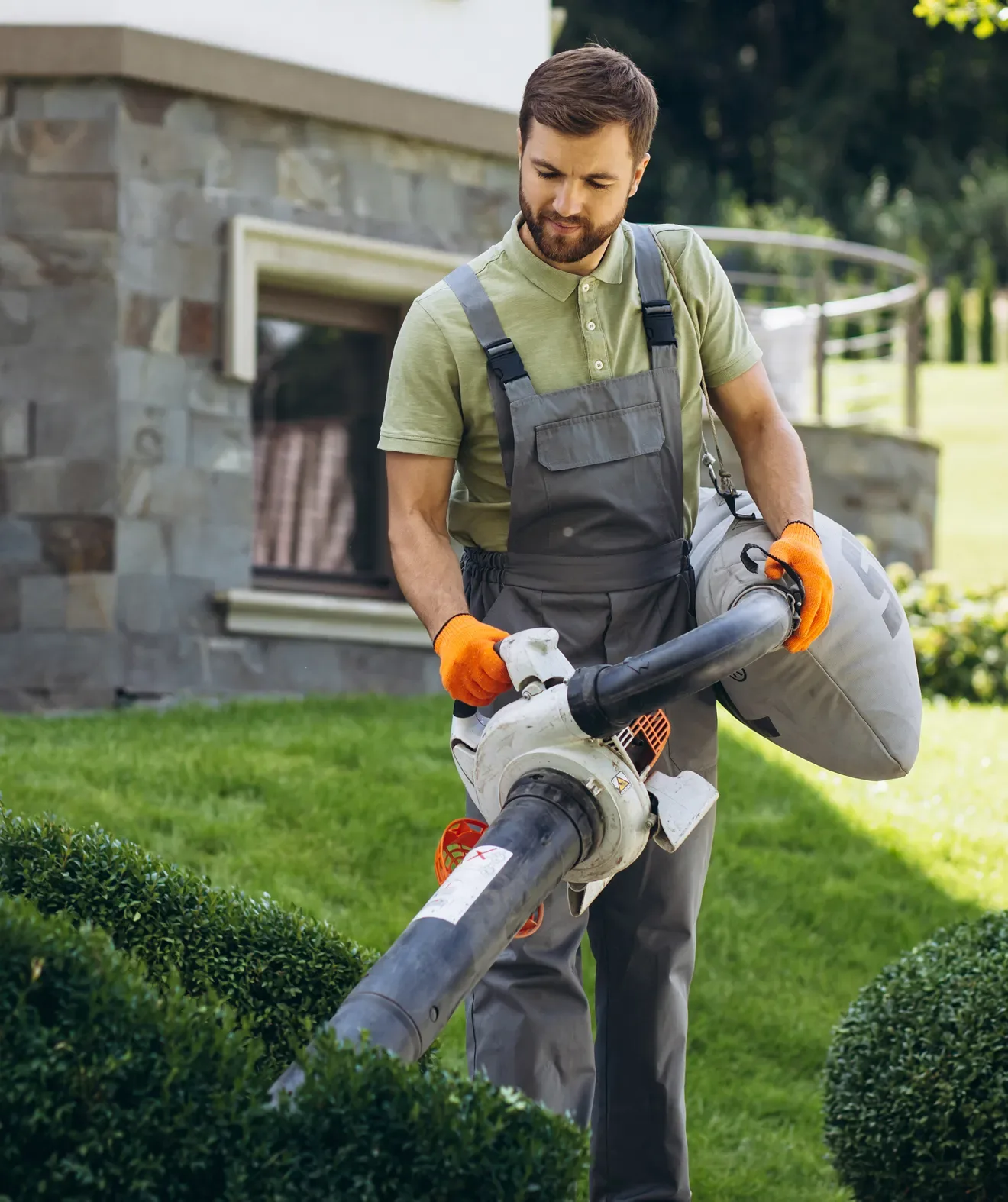 Man in work clothes using a leaf blower to clear bushes outside a stone house.