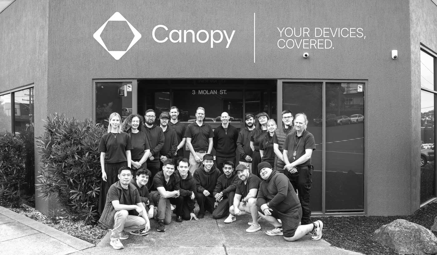Group of 19 people posing outside a building with Canopy logo and slogan, Your Devices Covered.