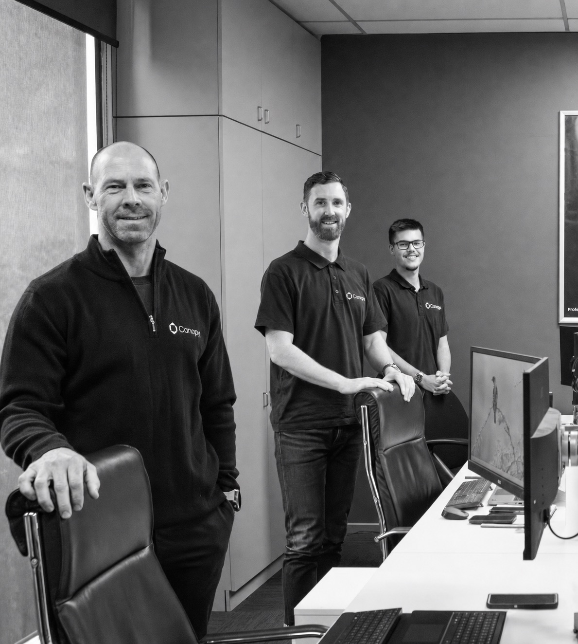 Three men in black Canopy shirts standing behind office chairs at a workstation with monitors.