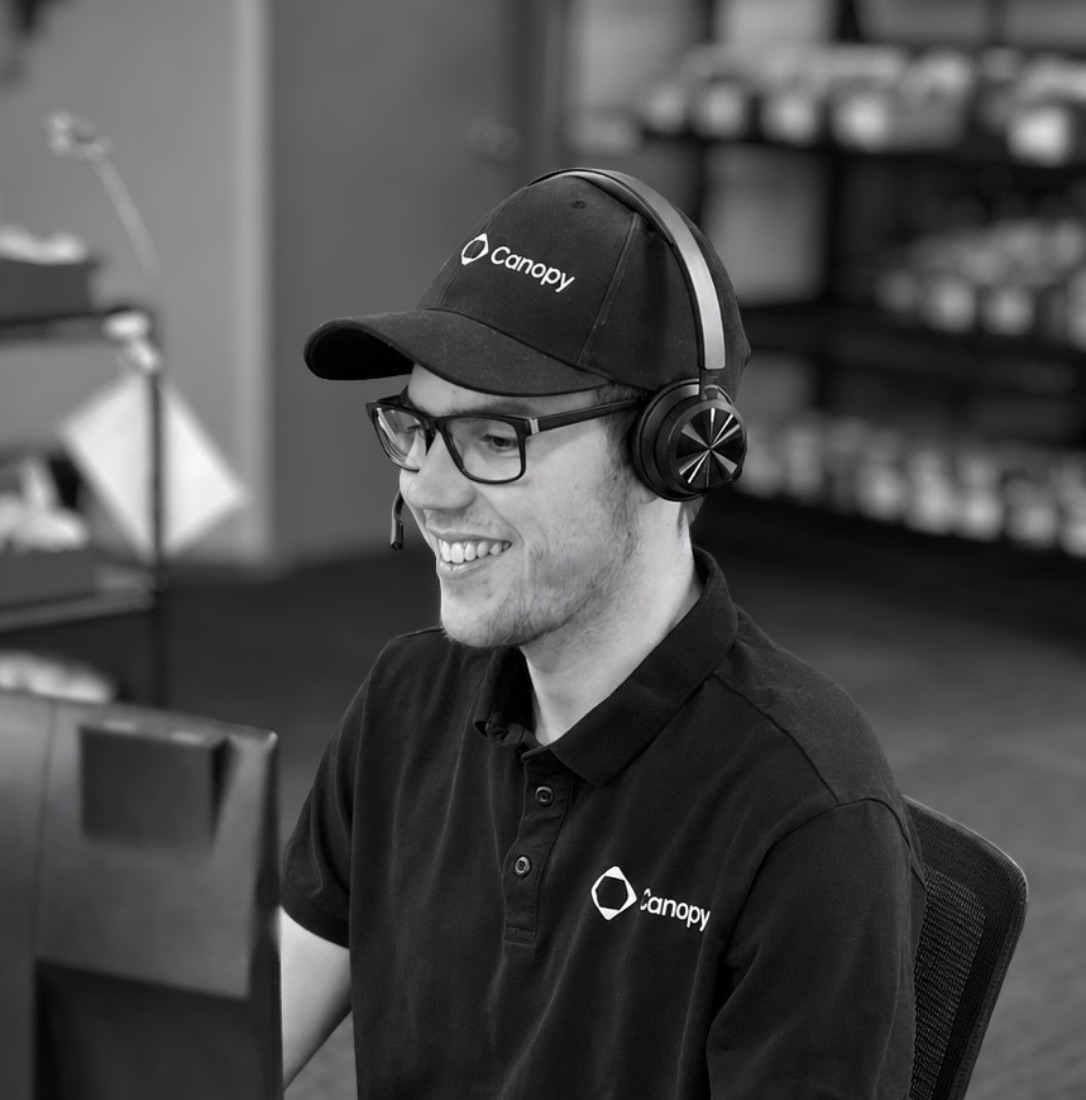 Smiling man wearing glasses, Canopy cap, polo, and headset, seated at a desk in an office.