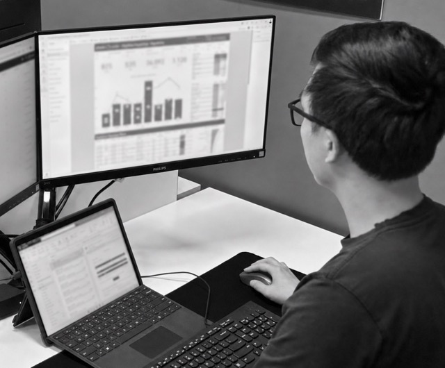 Person working at a desk with dual monitors and a laptop displaying charts and data.