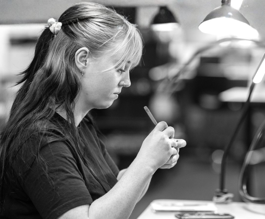 Woman with a ponytail working intently on a small object at a desk under a lamp.
