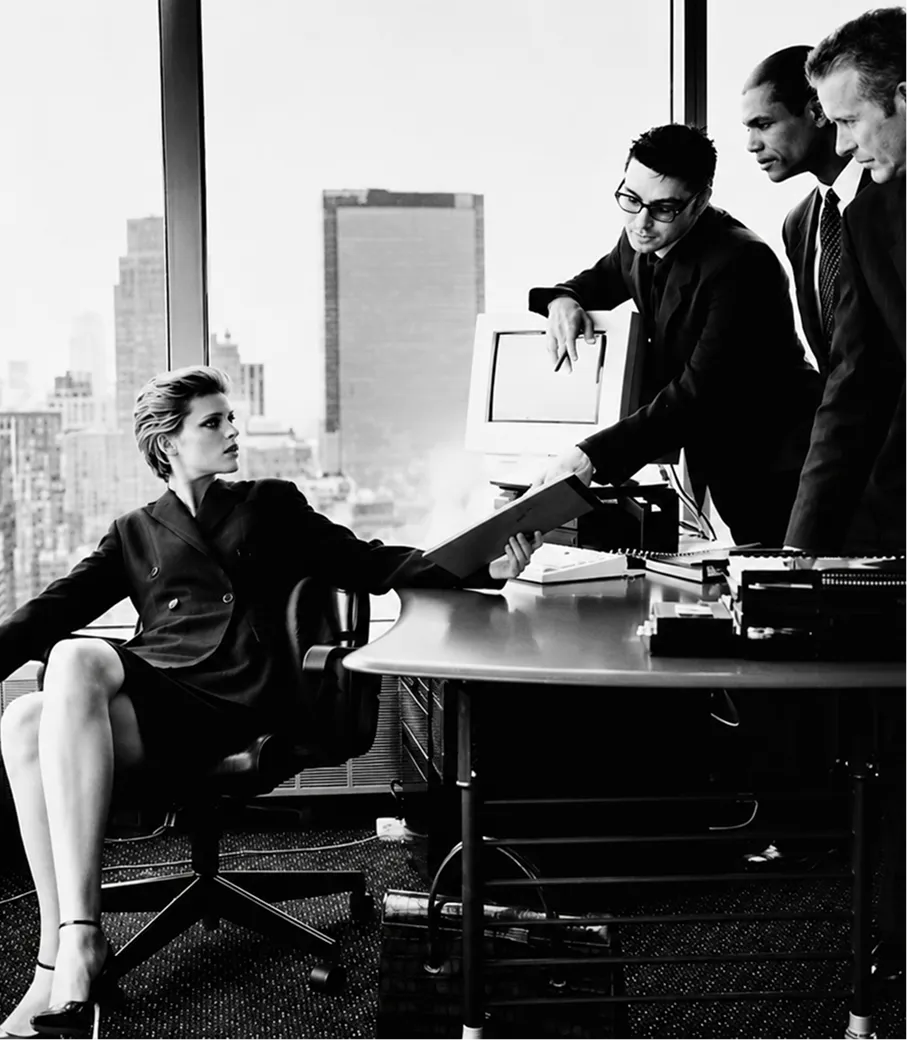Black and white photo of a woman sitting and three men standing at a desk in an office with city view.