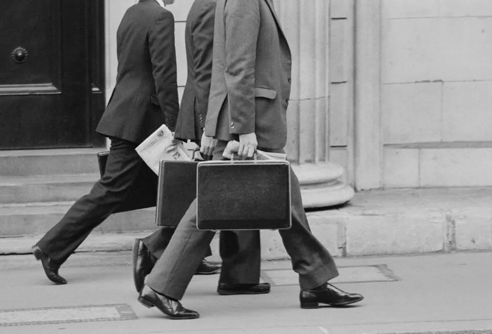 Three businessmen walking side by side carrying briefcases and a newspaper in black and white.