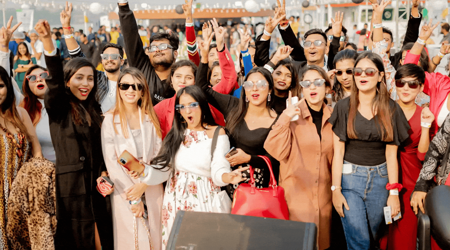 Group of young people at an outdoor event, many smiling and raising hands with peace signs, wearing sunglasses and casual to semi-formal clothing, appearing cheerful and energetic.