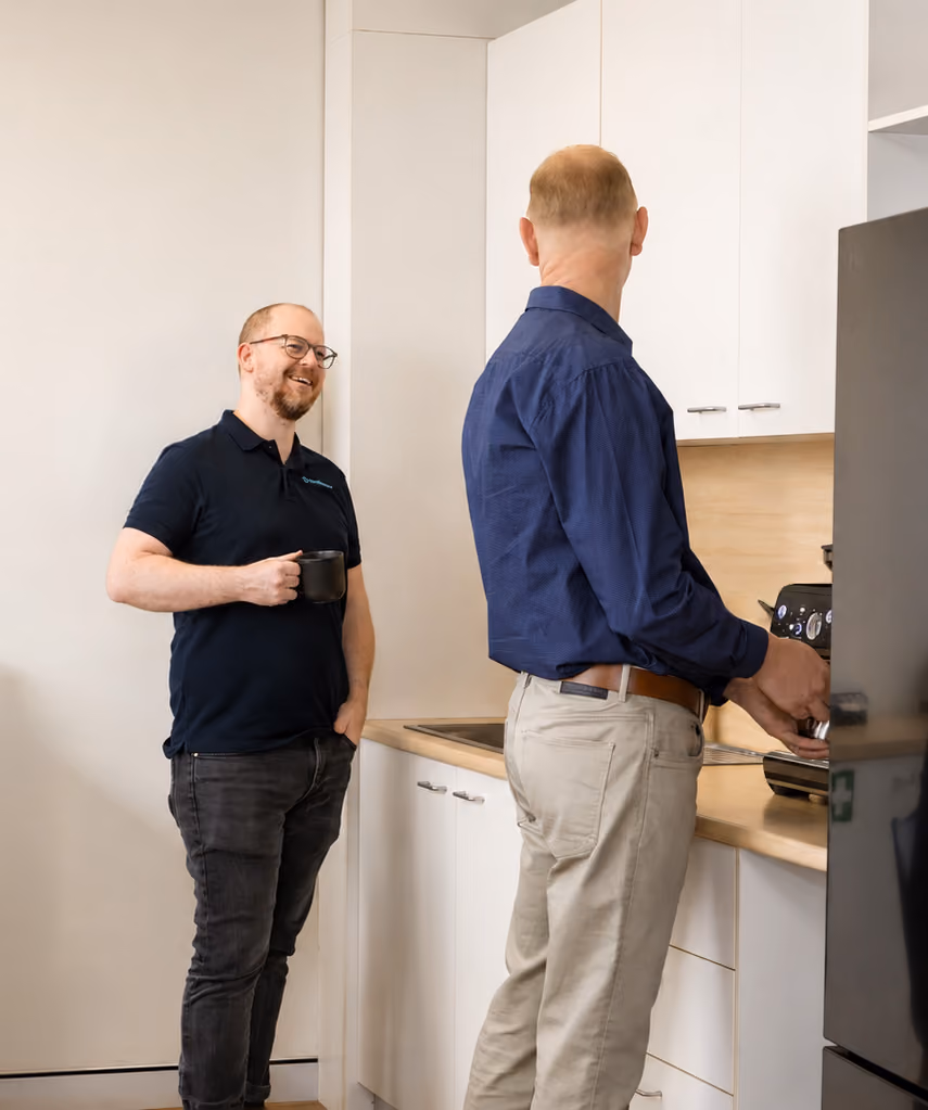 Two cyber team members chatting in the office kitchen; one holds a coffee cup while the other uses a coffee machine.